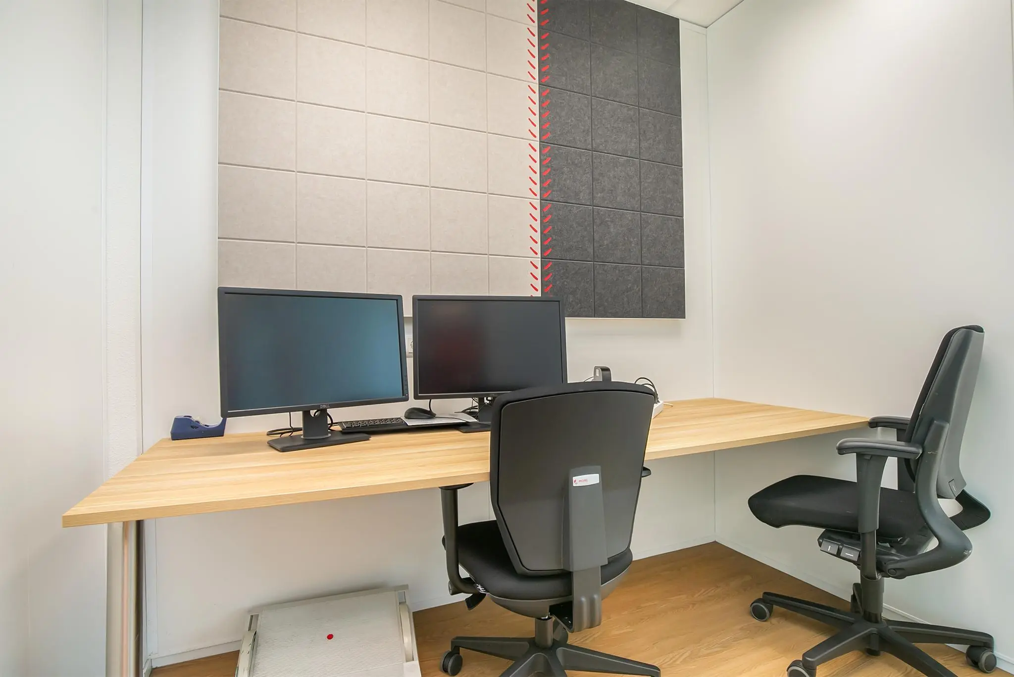 Kantoorruimte met twee bureaustoelen, twee beeldschermen op een houten bureau en geluidsabsorberende panelen aan de muur.