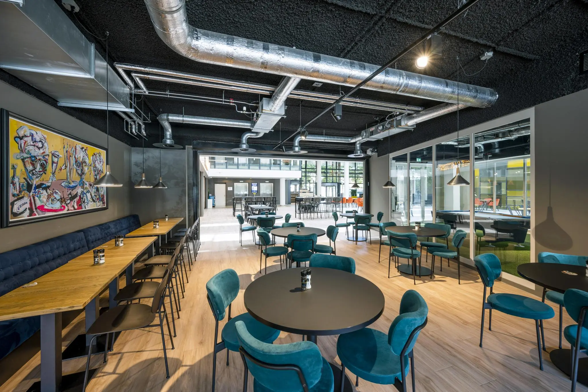 Moderne en kleurrijke horecaruimte aan de Thomas R. Malthusstraat met turquoise stoelen, houten vloeren en industriële plafondbuizen.