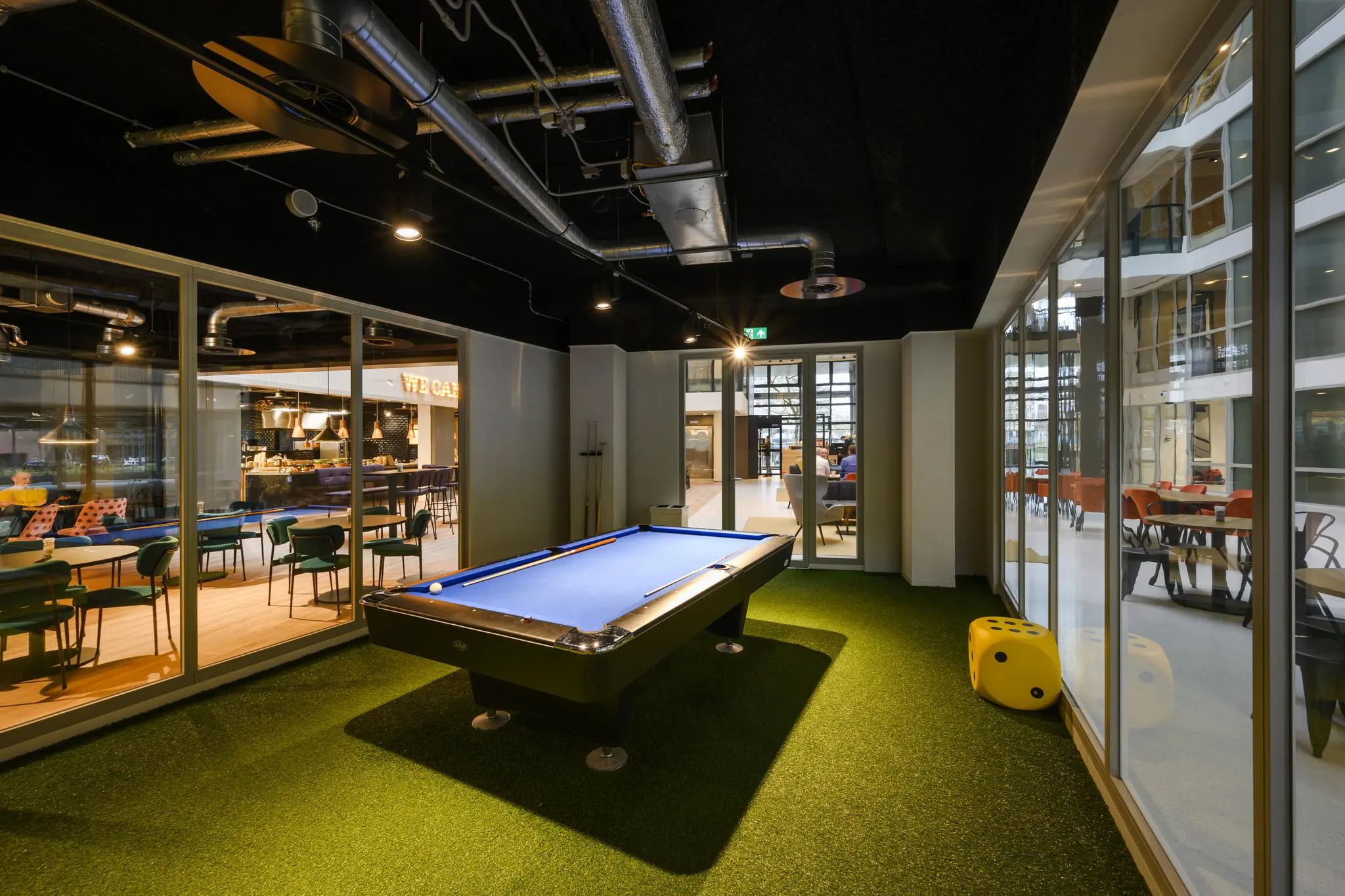 Moderne recreatieruimte aan de Thomas R. Malthusstraat met een pooltafel, kunstgrasvloer en zicht op een caféruimte.