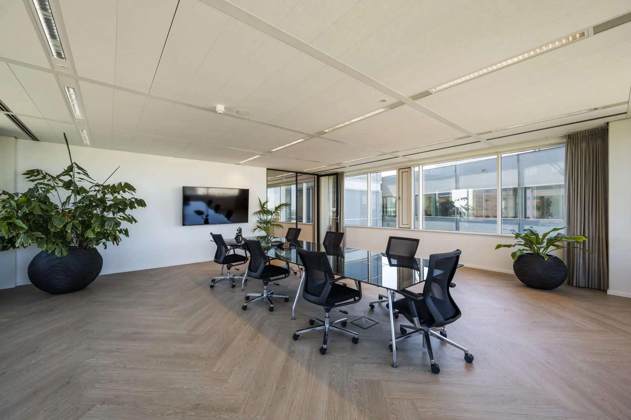 Moderne vergaderruimte aan de Thomas R. Malthusstraat met glazen tafel, zwarte stoelen, grote kamerplanten en een flatscreen aan de muur.
