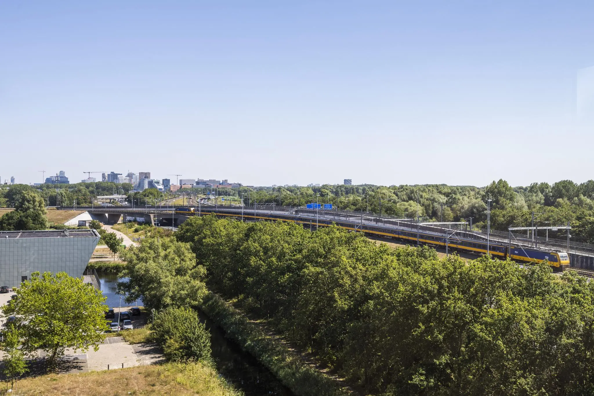 Panoramisch uitzicht op een trein die over het spoor rijdt bij de Thomas R. Malthusstraat in Amsterdam, omgeven door groen en stedelijke bebouwing in de verte.