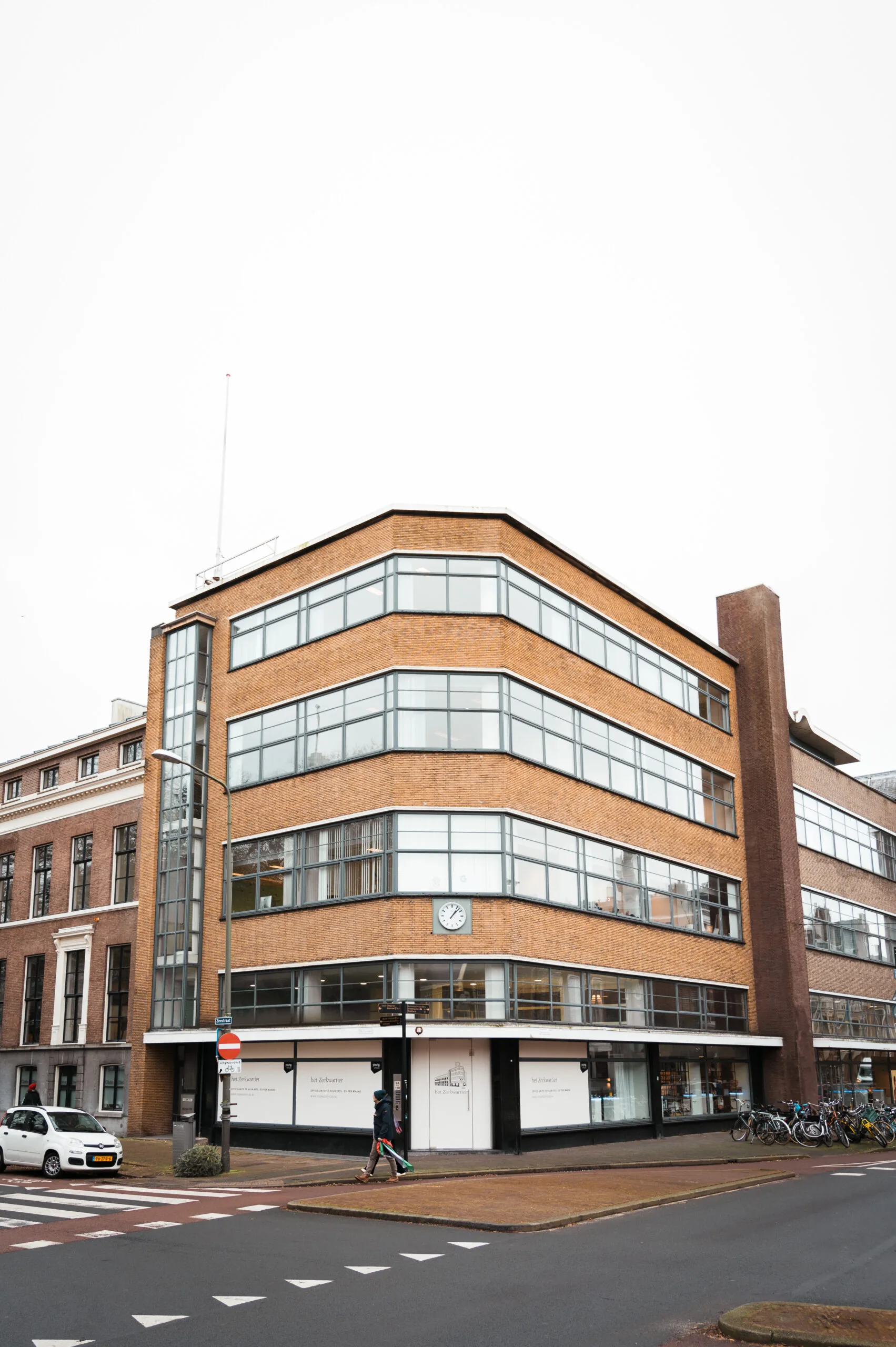 Bakstenen kantoorgebouw De Infirmerie op een straathoek met grote ramen en een klok aan de gevel.