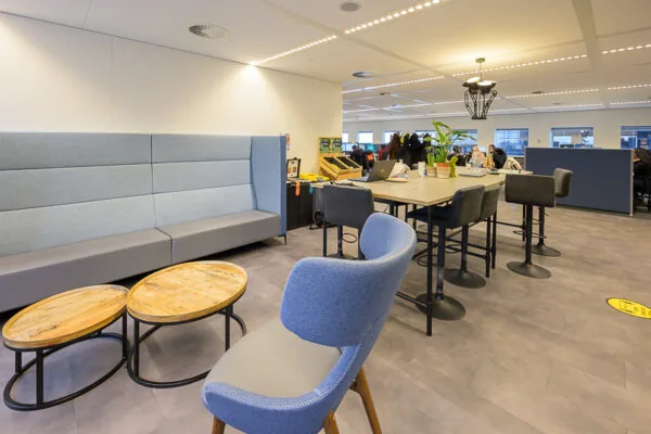Moderne kantoortuin met open werkplekken, een hoge vergadertafel en een zithoek met ronde houten tafels.