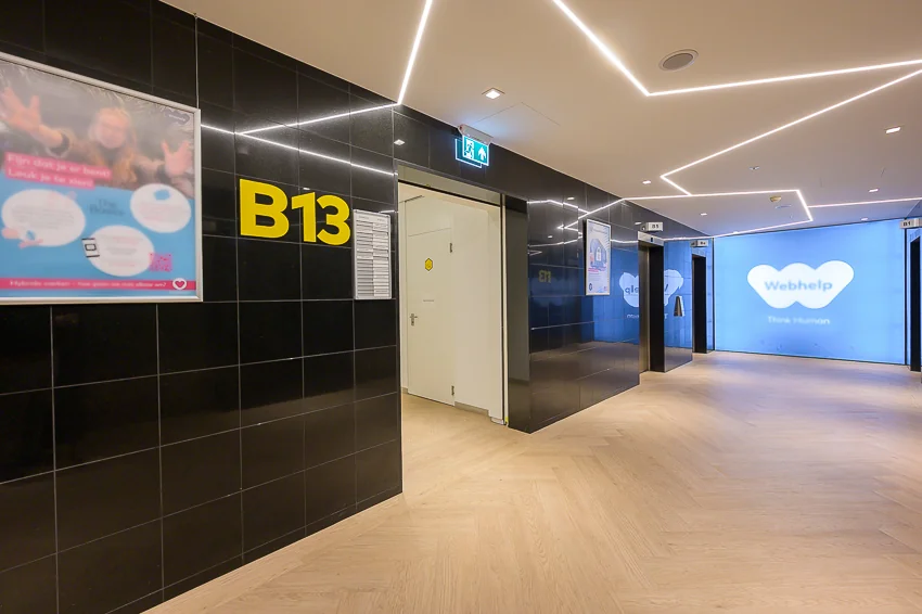 Moderne kantoorhal met liftdeuren, zwart betegelde muren met aanduiding B13, en een groot verlicht Webhelp-logo op de achtergrond.