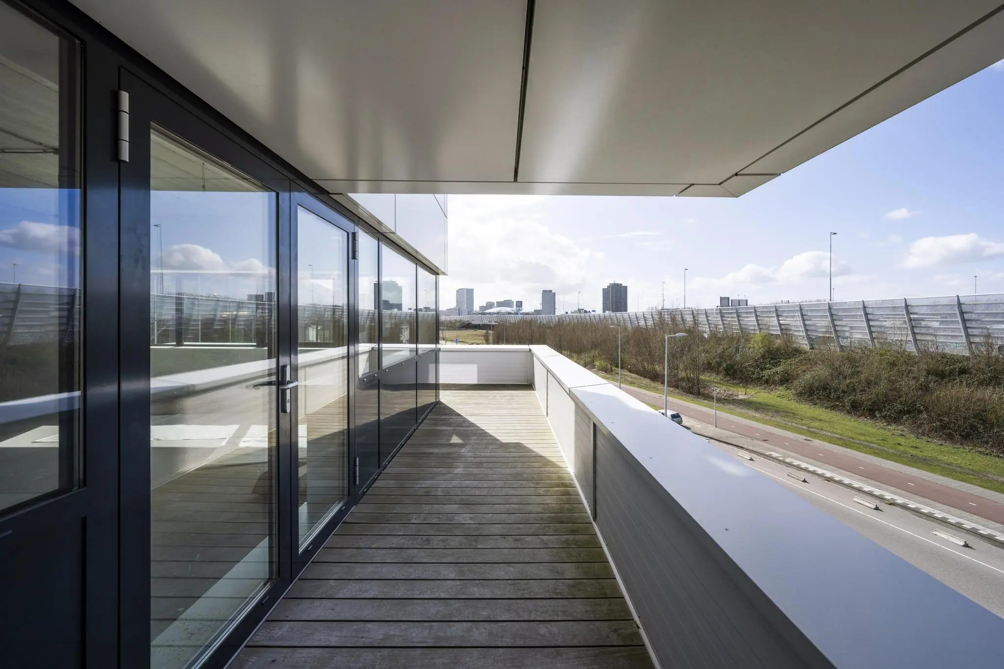 Modern balkon aan de Dynamostraat met houten vlonders en uitzicht op een geluidswal en hoogbouw in de verte.