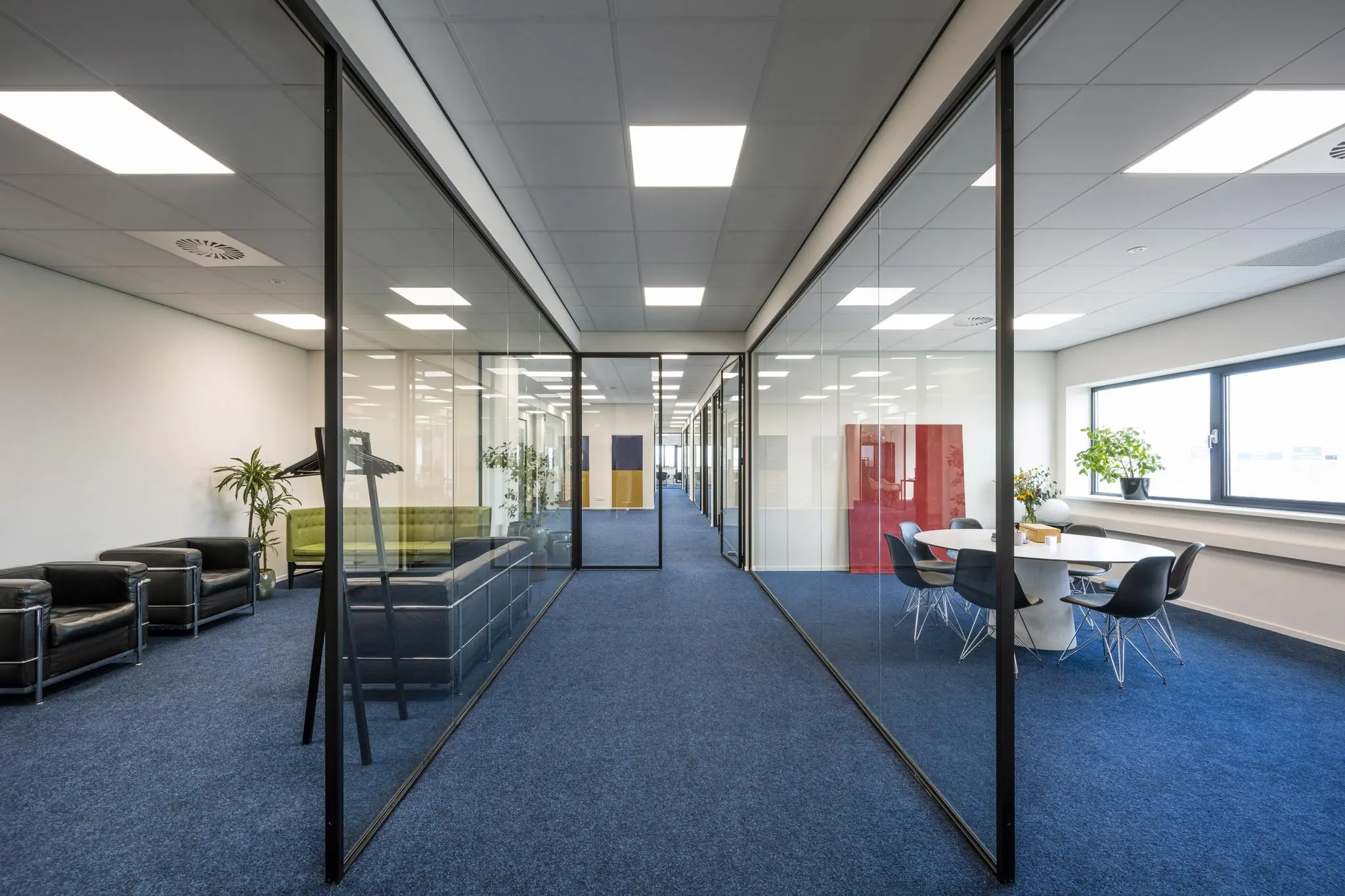 Moderne kantoorgang aan de Dynamostraat met glazen wanden, vergaderruimte en wachtruimte met stoelen en planten.