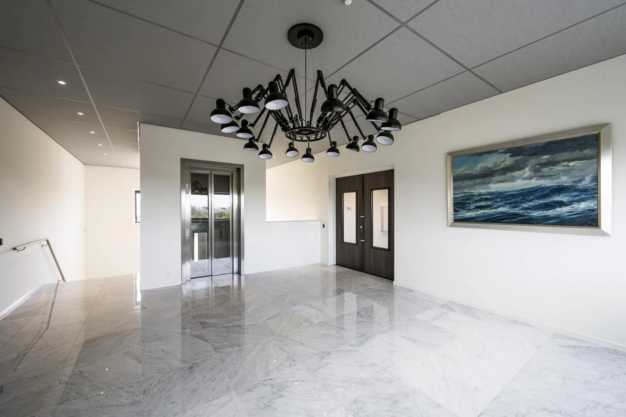 Moderne, lichte entreehal aan de Dynamostraat met marmeren vloer, opvallende zwarte plafondlamp, lift en schilderij van een woeste zee.