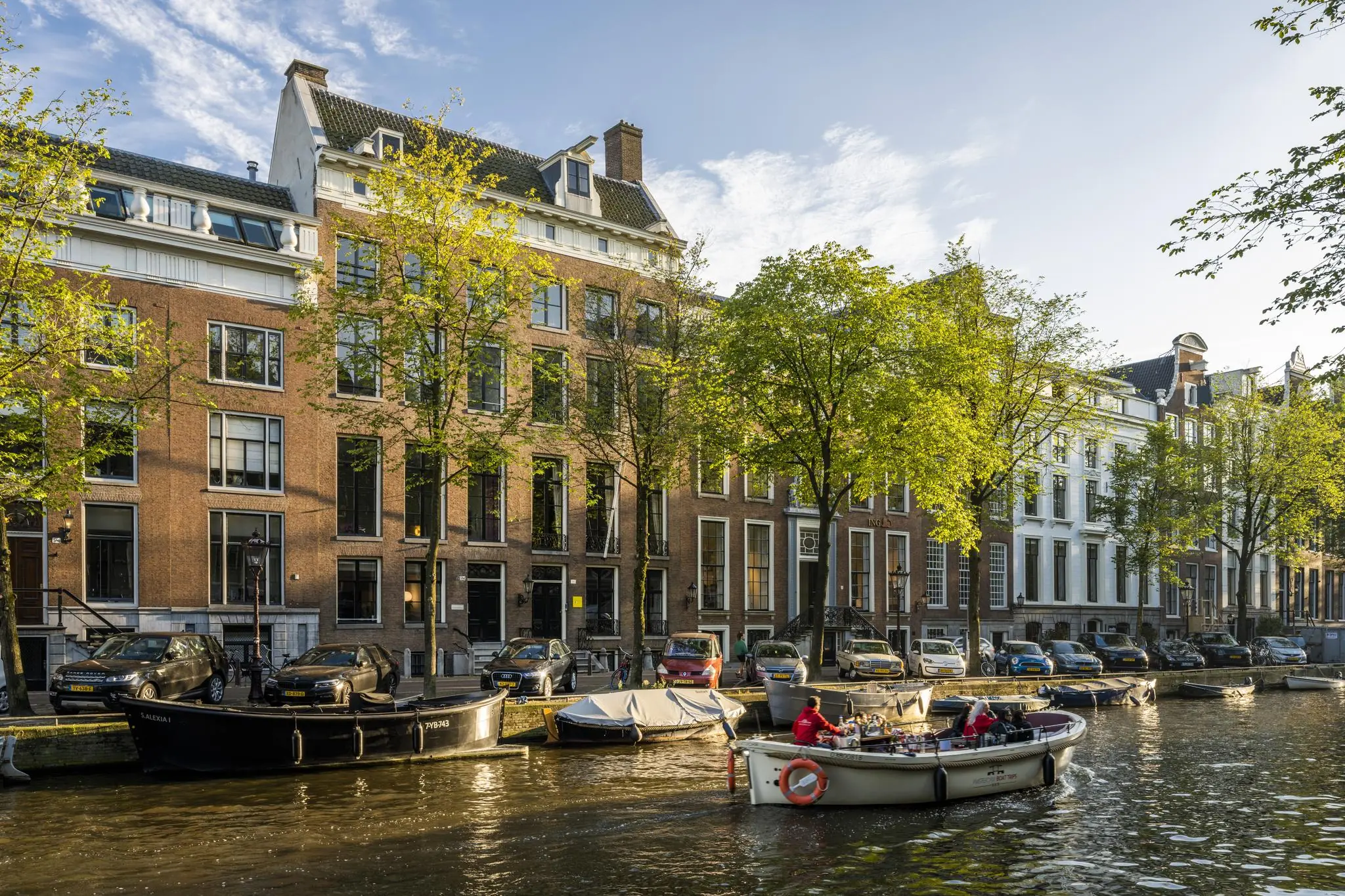 Zicht op de Herengracht in Amsterdam met woonboten, auto's en historische grachtenpanden in de avondzon.