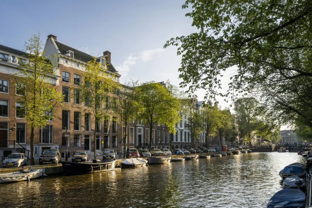 Gracht met woonboten, geparkeerde auto’s en historische panden langs de Herengracht in Amsterdam bij zonnig weer.