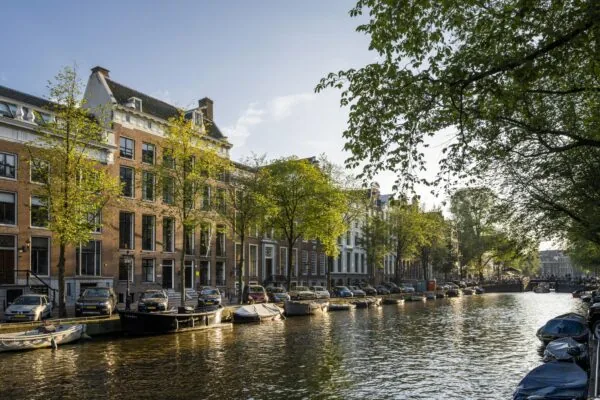 Gracht met woonboten, geparkeerde auto’s en historische panden langs de Herengracht in Amsterdam bij zonnig weer.