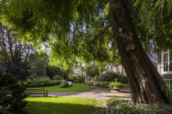 Groene binnentuin aan de Herengracht met grote bomen, kunstobjecten en bankjes in de schaduw.