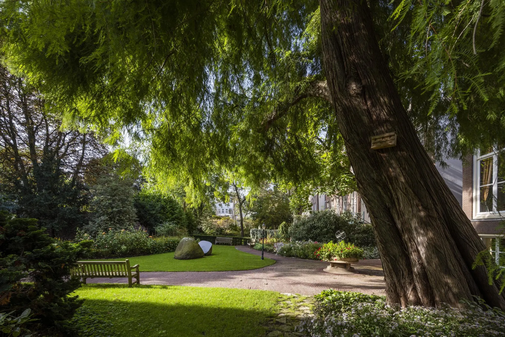 Groene binnentuin aan de Herengracht met grote bomen, kunstobjecten en bankjes in de schaduw.