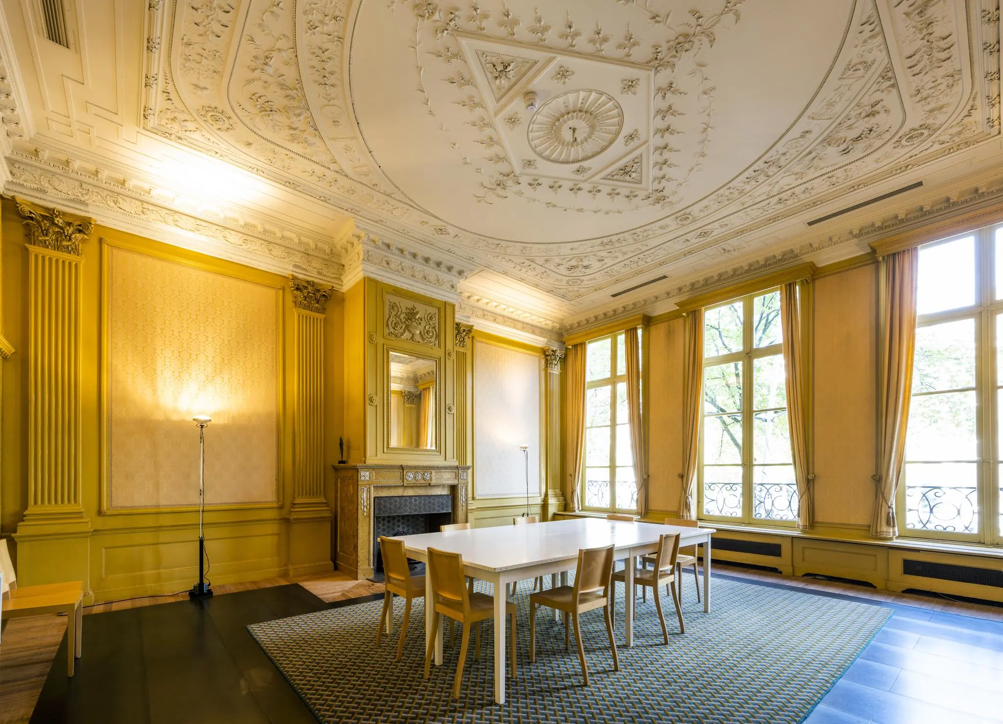 Elegante vergaderruimte aan de Herengracht met hoge ramen, goudgele wanden, een rijk gedecoreerd plafond en een moderne tafel met stoelen.
