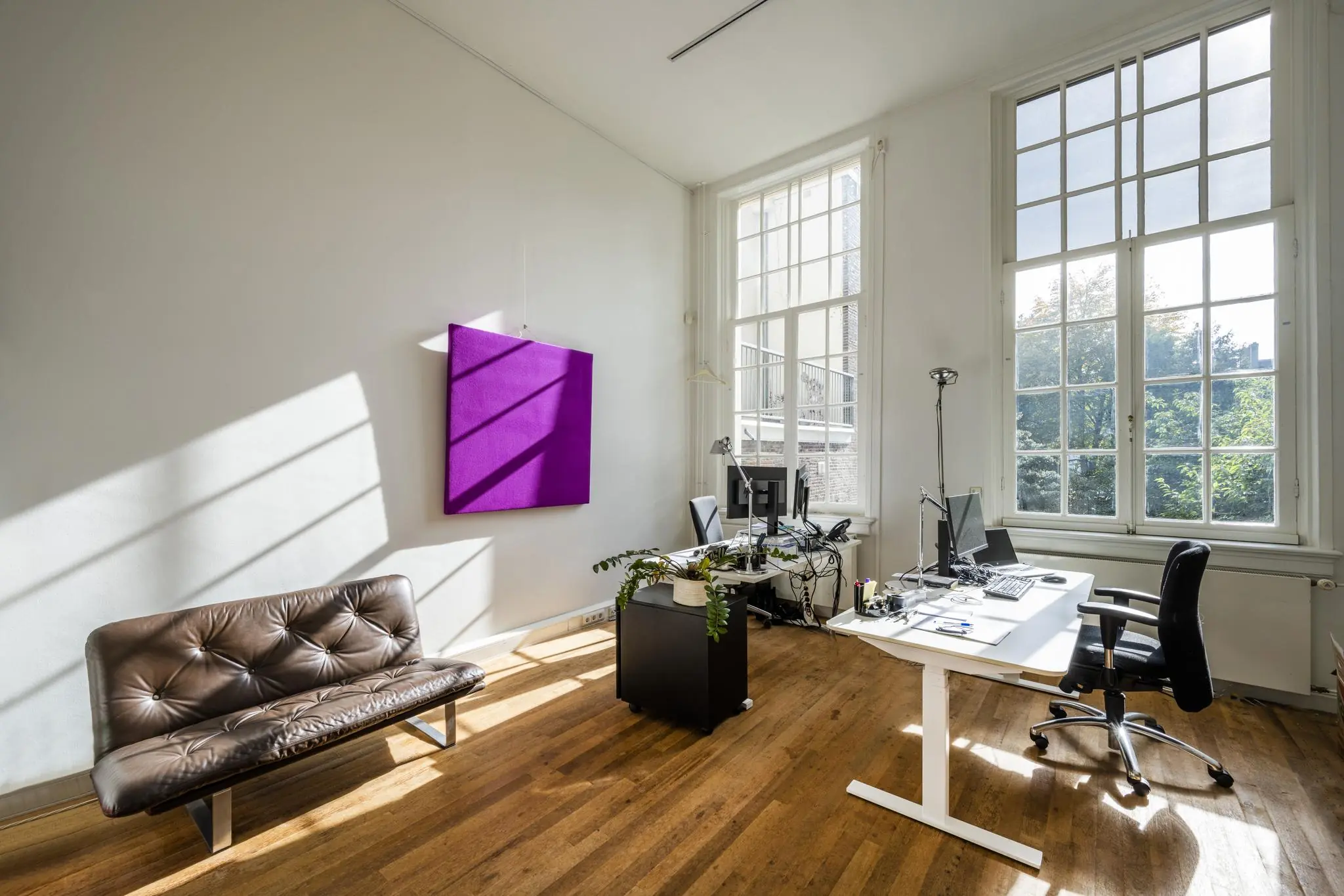 Licht en modern kantoor aan de Herengracht met hoge ramen, een bureauopstelling en een paarse wanddecoratie.