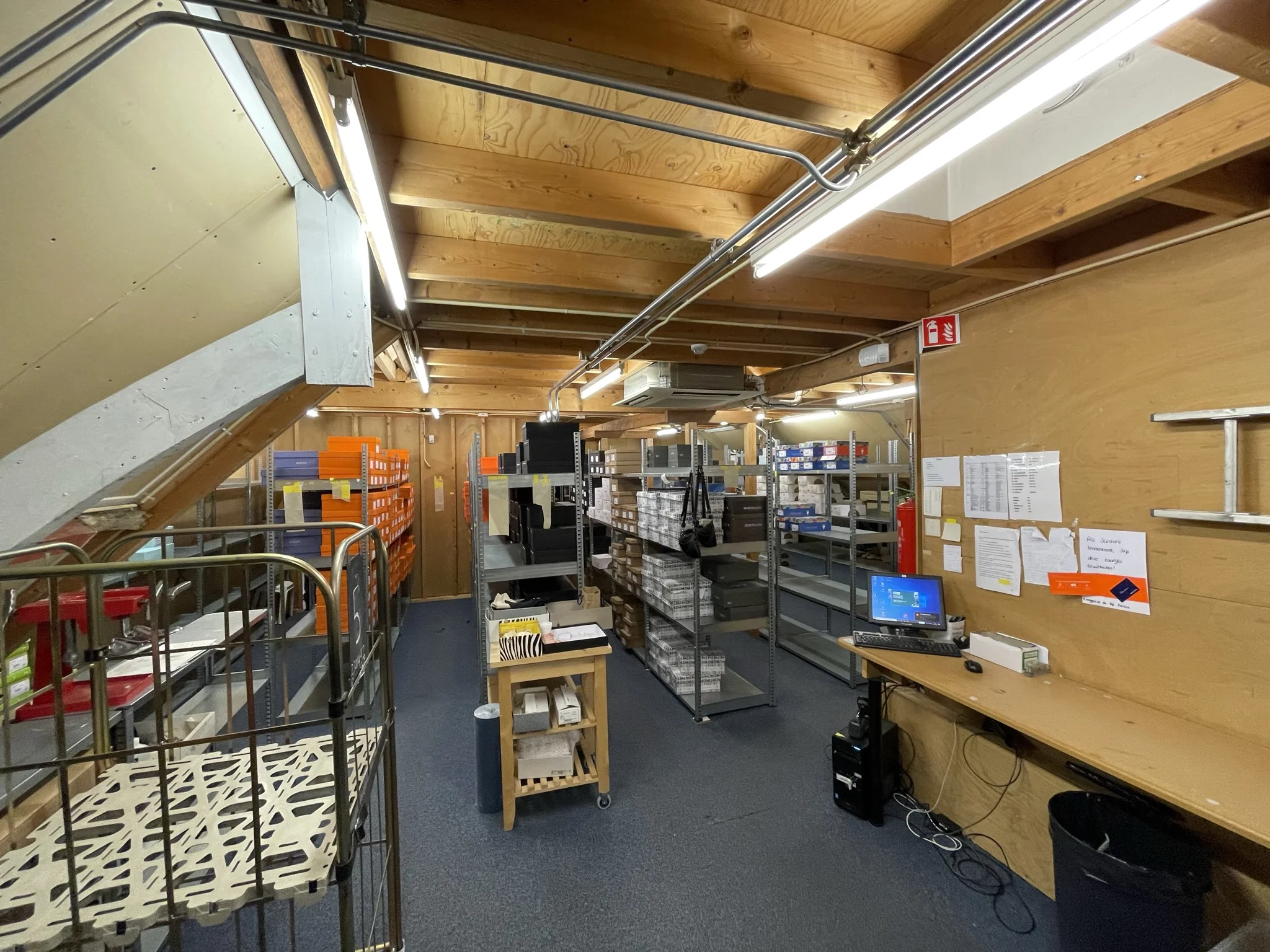 Magazijnruimte met stellingen vol schoenendozen, een werkplek met computer en houten plafondconstructie.