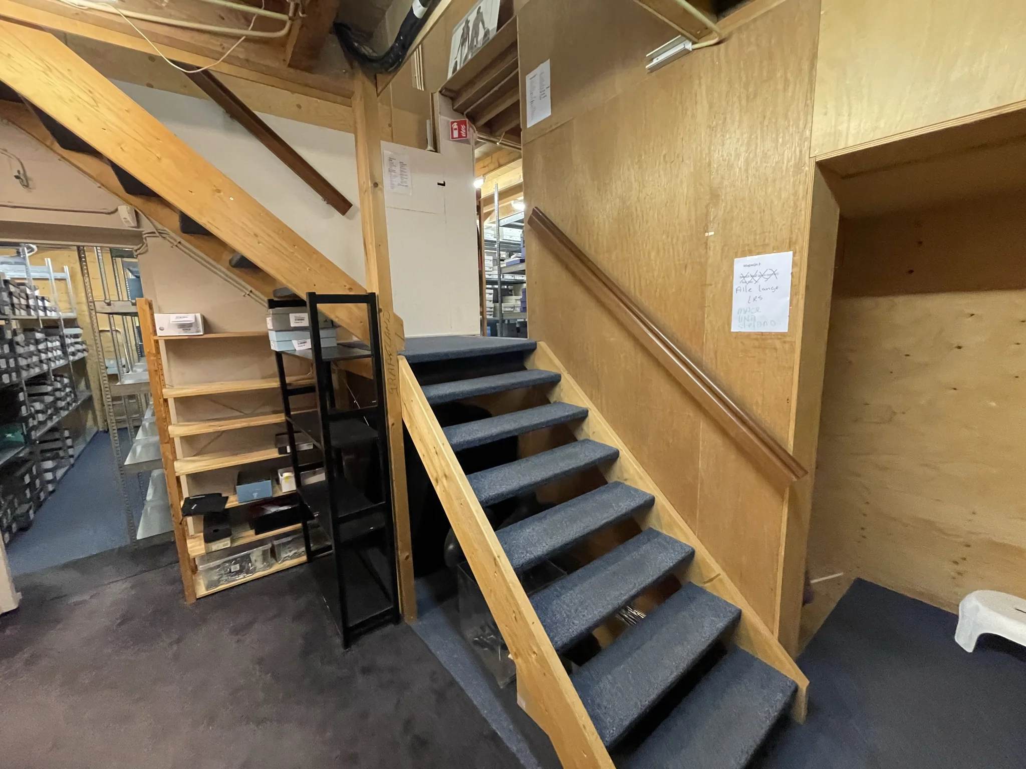 Trapopgang met houten leuningen en tapijt in een opslagruimte aan de Molendijk, omgeven door rekken met dozen en materialen.