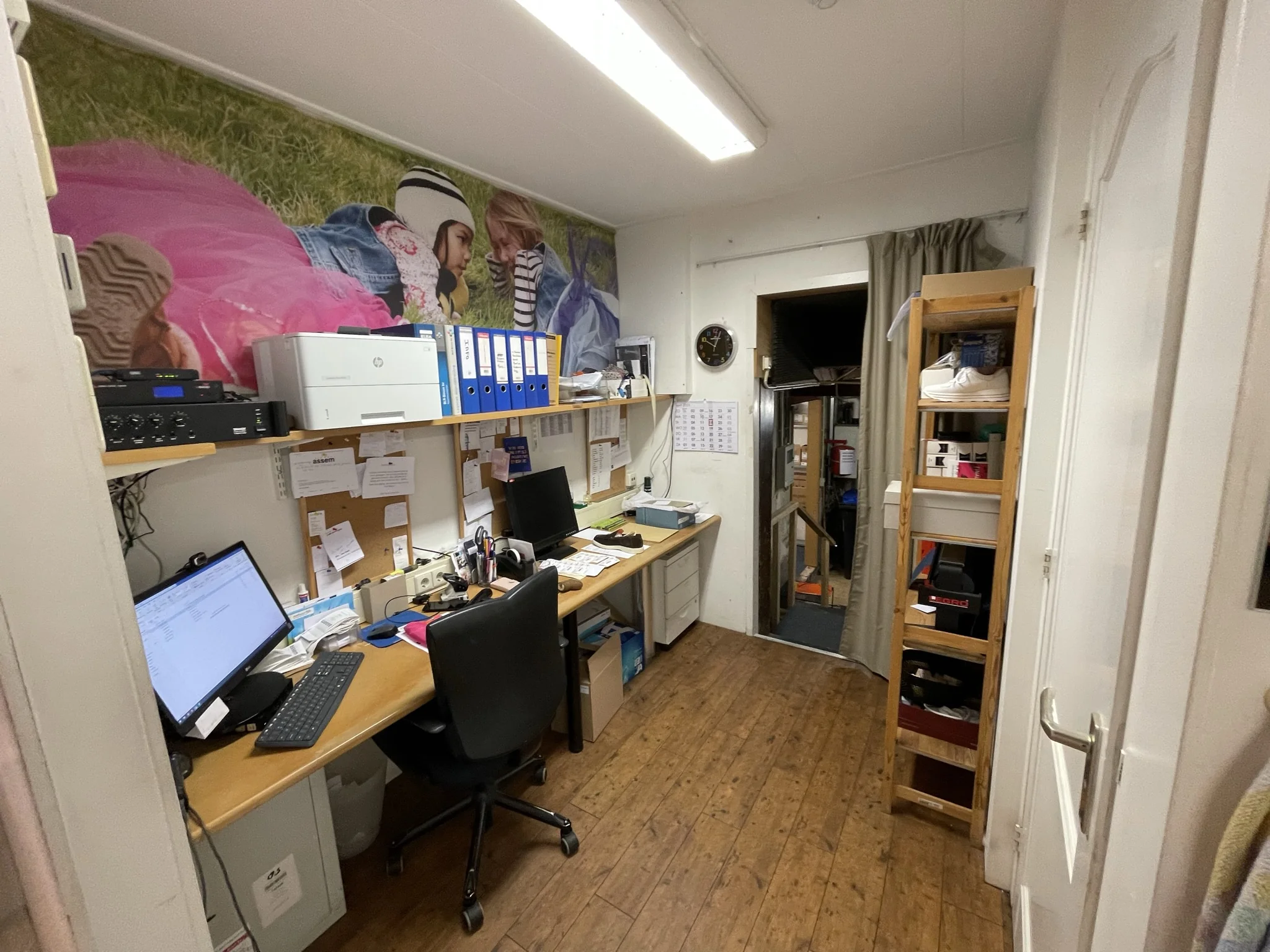 Kleine kantoorruimte met twee werkplekken, houten vloer, opbergrekken en een grote muurfoto van spelende kinderen aan de wand.