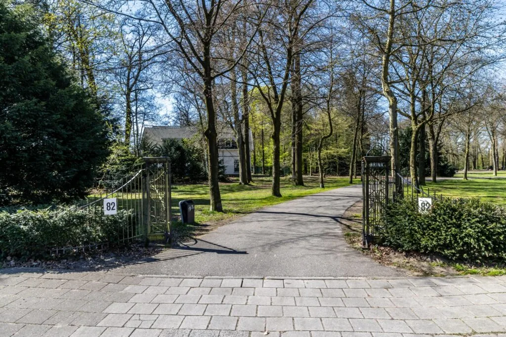 Oprit met sierhek en huisnummer 82 aan de Utrechtseweg, omgeven door bomen en groen.