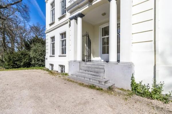 Ingang met trap en zuilen van een monumentaal pand aan de Utrechtseweg, omgeven door bomen en grindpad.