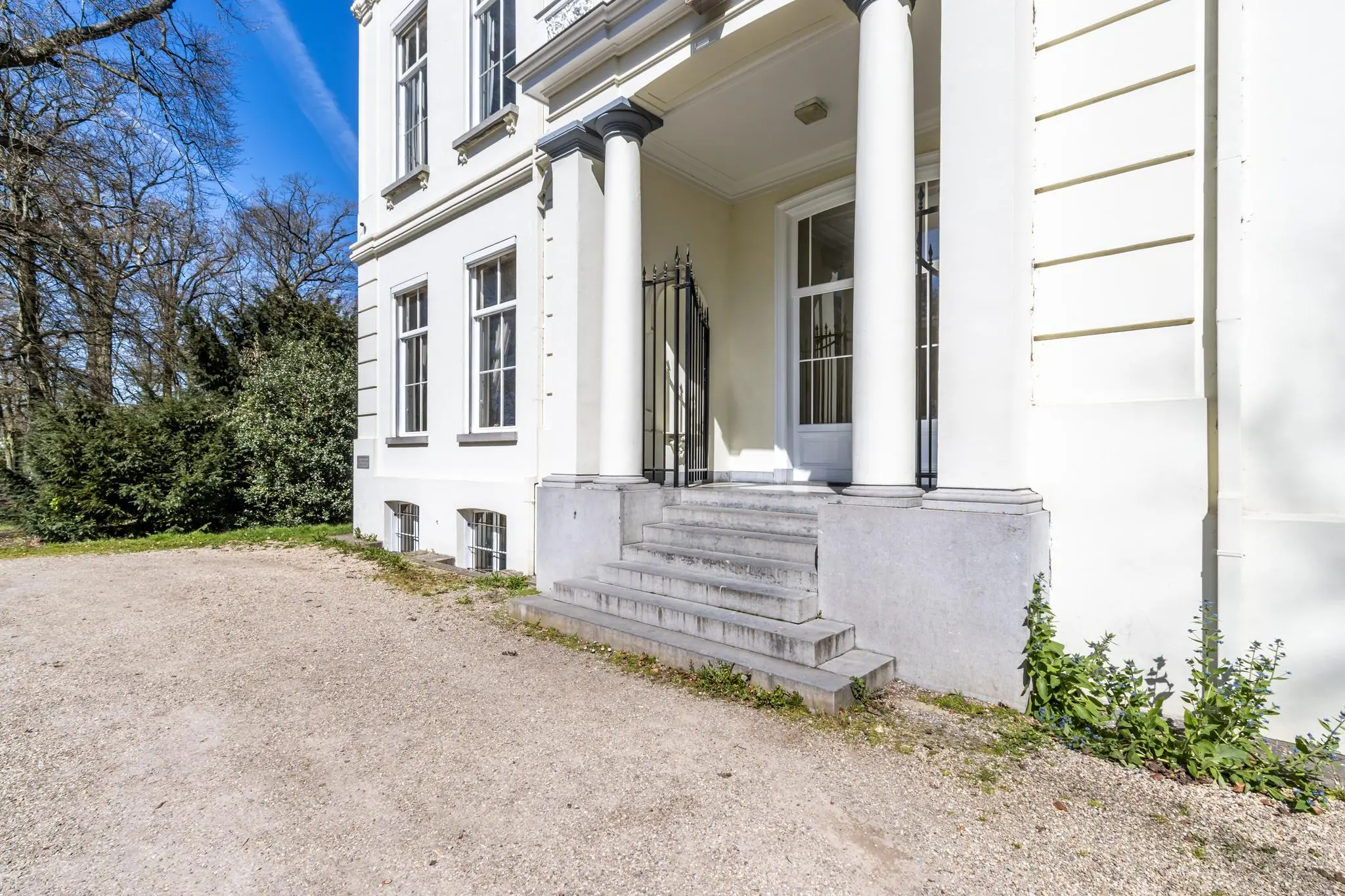 Ingang met trap en zuilen van een monumentaal pand aan de Utrechtseweg, omgeven door bomen en grindpad.