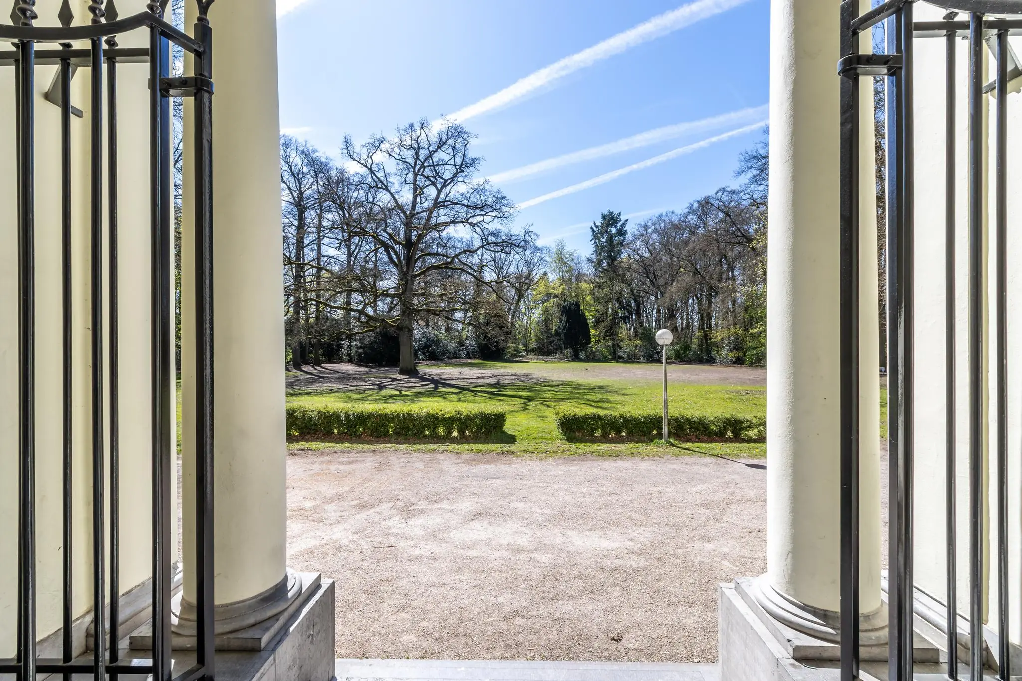 Uitzicht vanuit een klassiek portiek aan de Utrechtseweg op een zonnige tuin met grote bomen en een open grasveld.