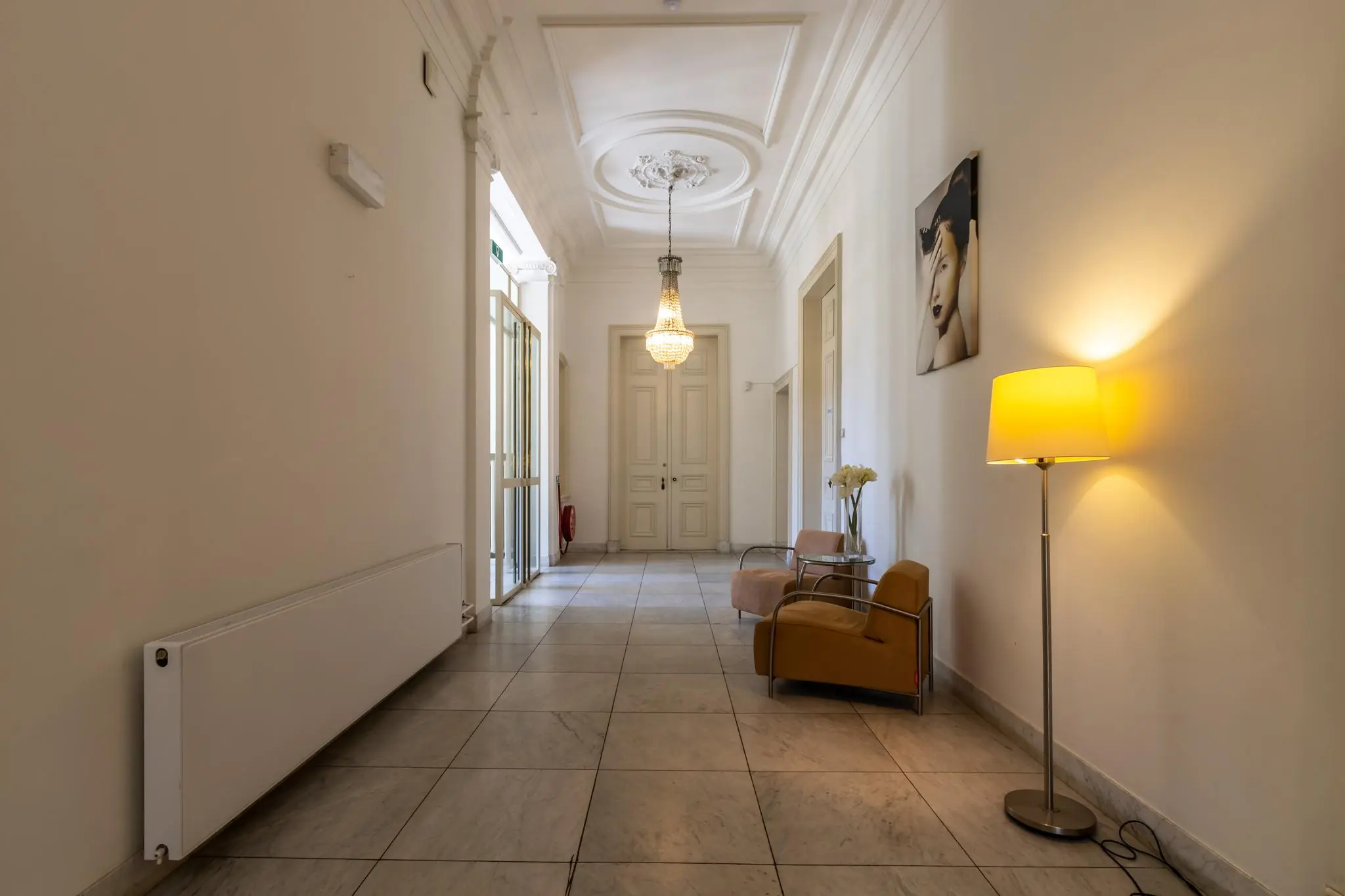 Elegante gang met marmeren vloer, klassieke deuren, een kroonluchter en een zithoek met vloerlamp en moderne kunst aan de muur.