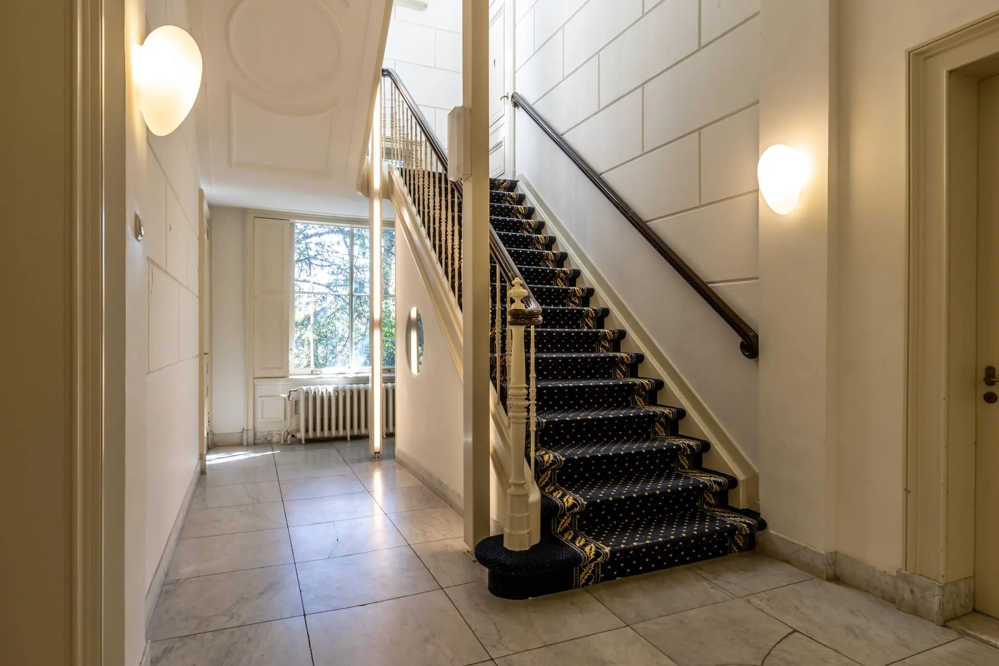 Trap met donkerblauw tapijt en marmeren vloer in een klassiek entreehal aan de Utrechtseweg.