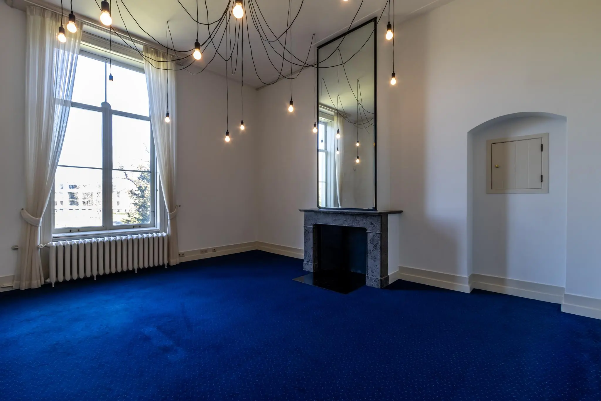 Lichte kamer aan de Utrechtseweg met blauw tapijt, hoge ramen, een marmeren schouw en moderne hangverlichting.