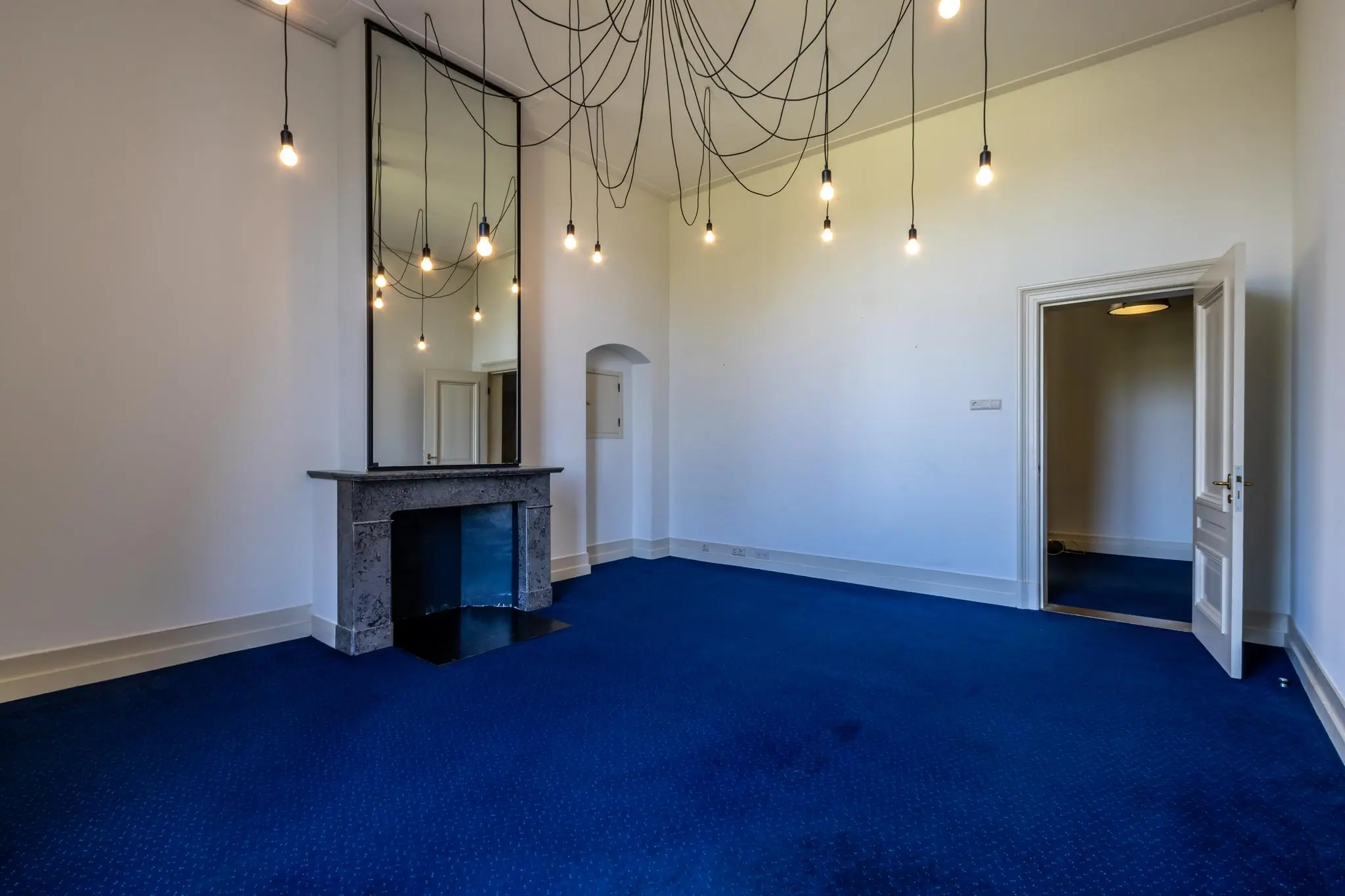 Ruime kamer aan de Utrechtseweg met blauwe vloerbedekking, een marmeren schouw, een grote spiegel en meerdere hanglampen aan het plafond.
