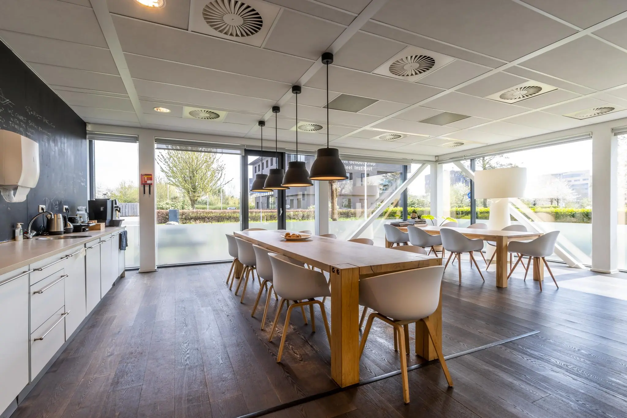 Moderne, lichte kantoorkeuken met houten tafels, witte stoelen en grote ramen aan de Groenewoudsedijk.