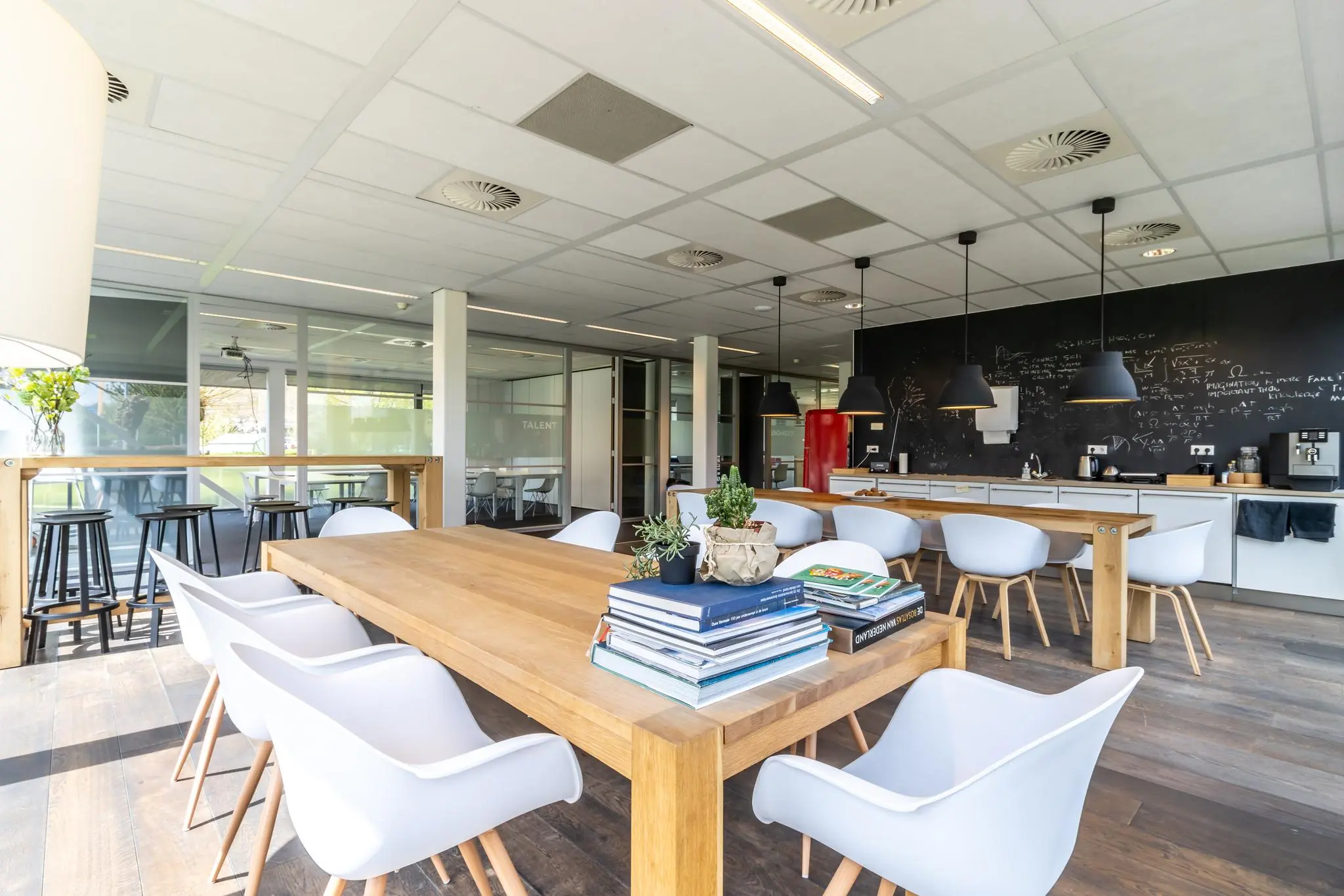 Moderne, lichte kantoorruimte aan de Groenewoudsedijk met een grote houten tafel, witte stoelen, boekstapel en een keuken met zwarte krijtbordmuur.