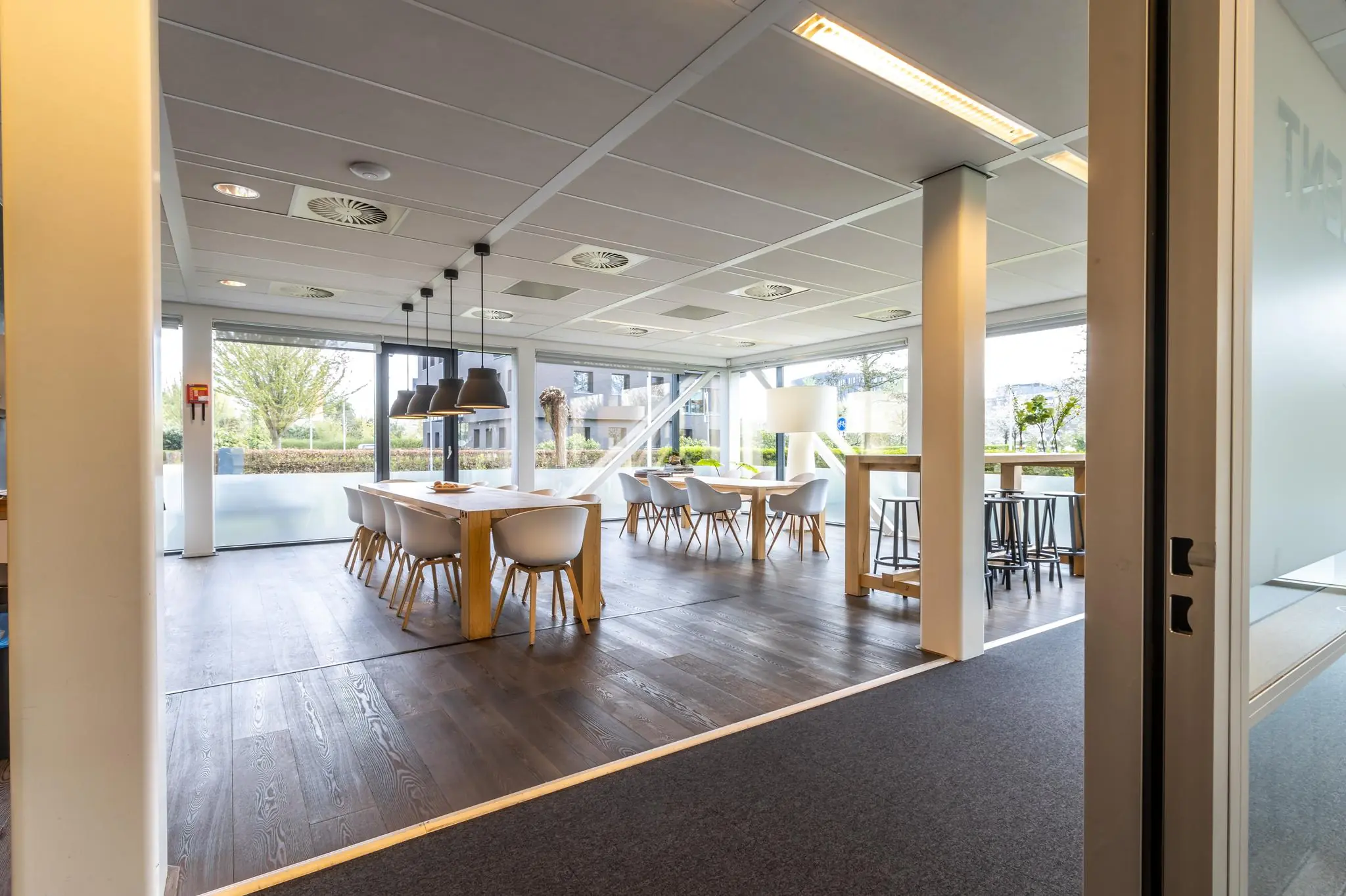 Moderne, lichte kantineruimte aan de Groenewoudsedijk met houten tafels, witte stoelen en grote ramen.