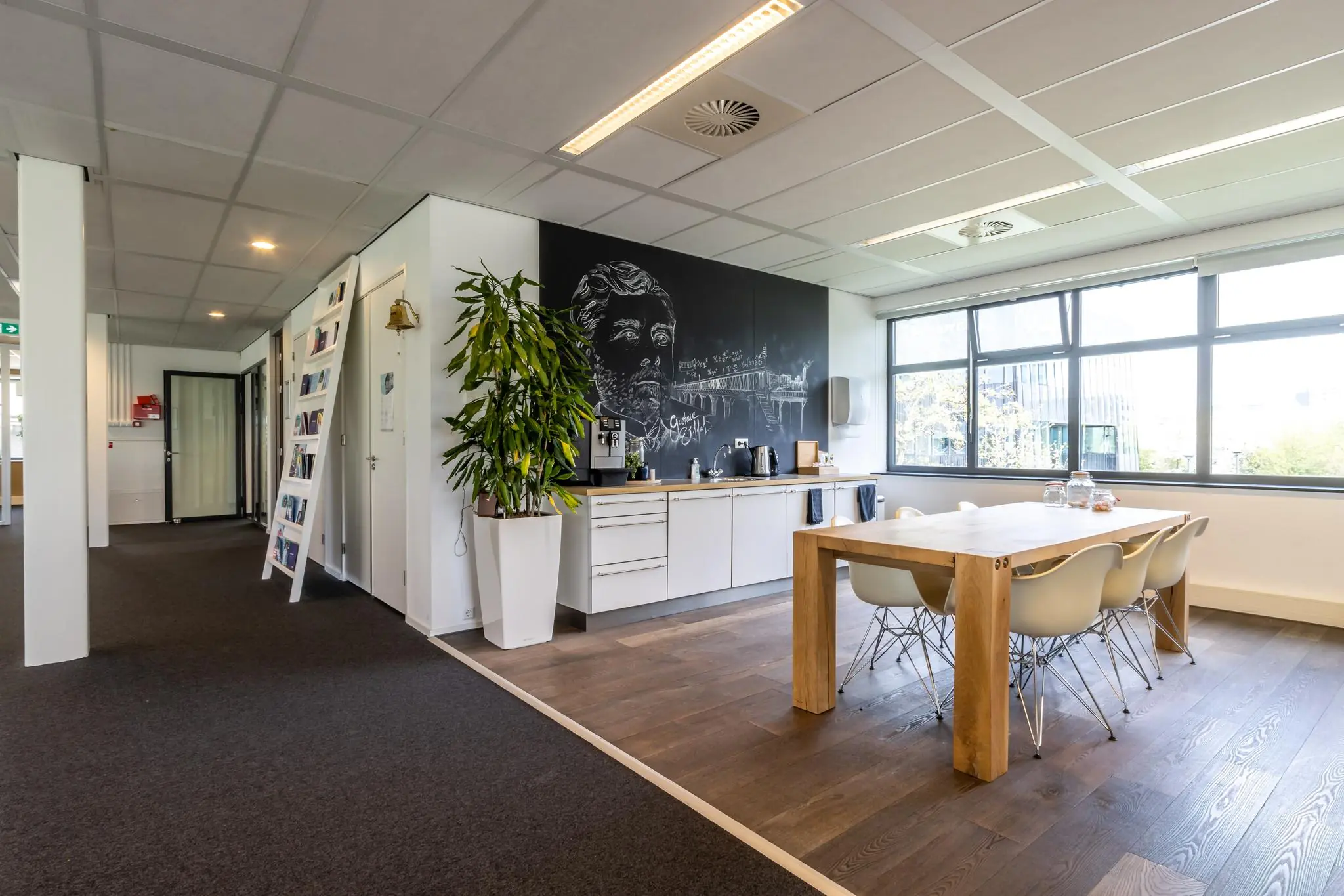 Moderne kantoorkeuken met houten eettafel, wit meubilair en een zwart schoolbord met krijttekening aan de muur.