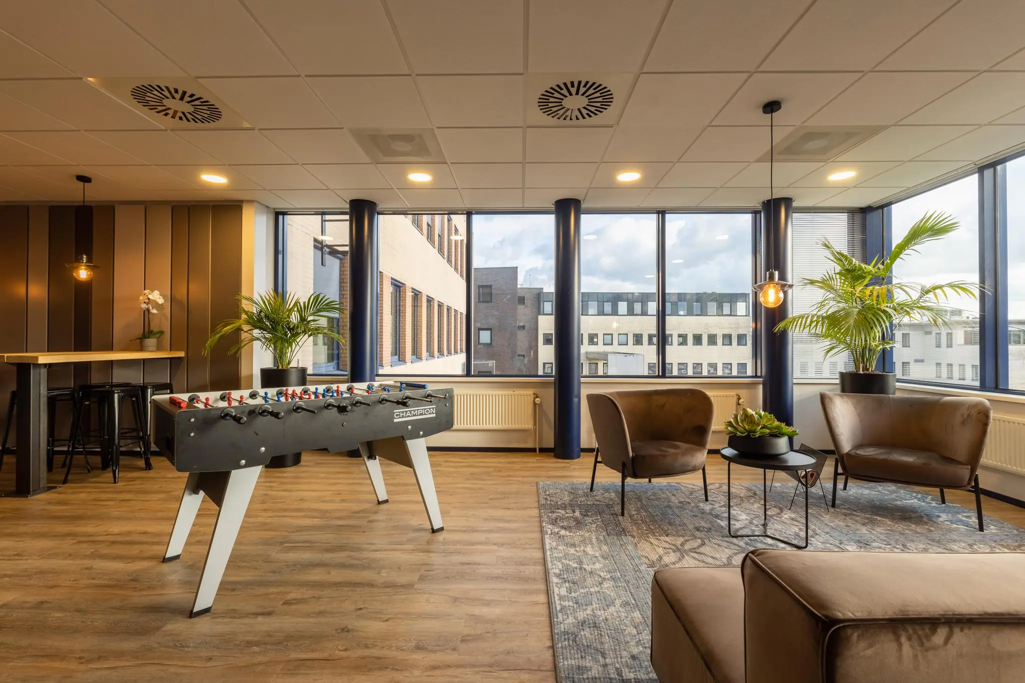 Moderne kantoorlounge aan de Fokkerstraat met tafelvoetbaltafel, zithoek en grote ramen met uitzicht op omliggende gebouwen.