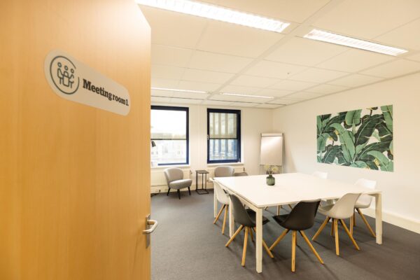 Vergaderruimte met moderne inrichting, grote tafel, diverse stoelen en muurkunst, gezien vanuit een openstaande deur met het bordje "Meetingroom 1".