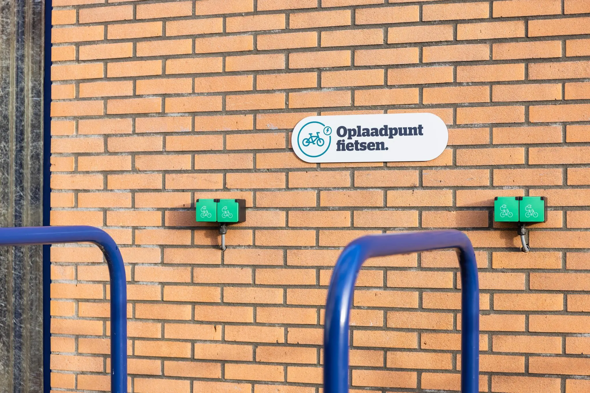 Oplaadpunt voor fietsen aan een bakstenen muur met twee groene laadstations en een bijbehorend bord.