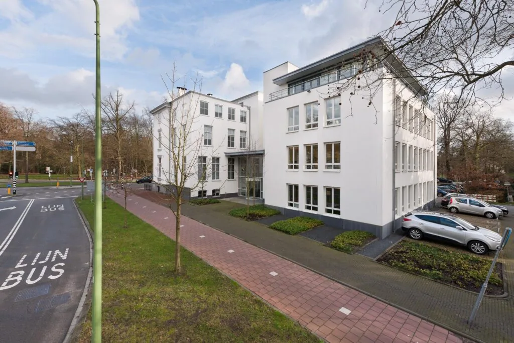 Wit kantoorgebouw aan de Driebergseweg met parkeerplaatsen en aangrenzende bushalte.