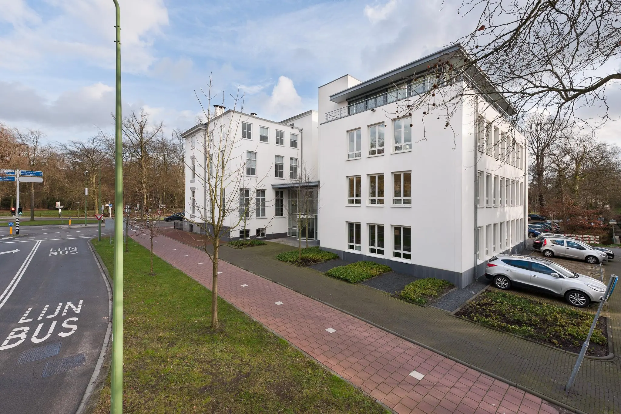 Wit kantoorgebouw aan de Driebergseweg met parkeerplaatsen en aangrenzende bushalte.