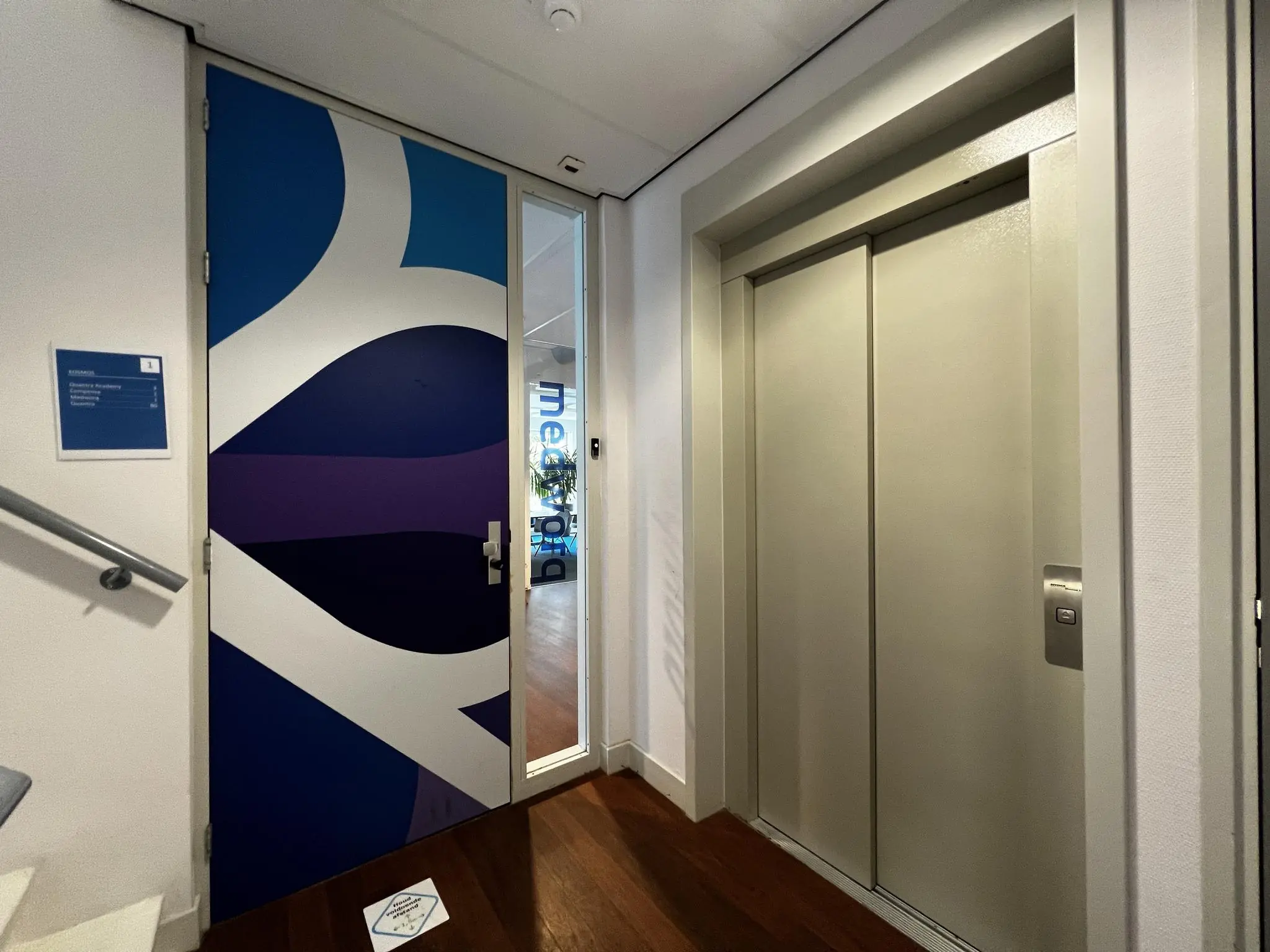 Ingang van een kantoorruimte aan de Driebergseweg, met een kleurrijke deur naast een gesloten liftdeur in een modern interieur.