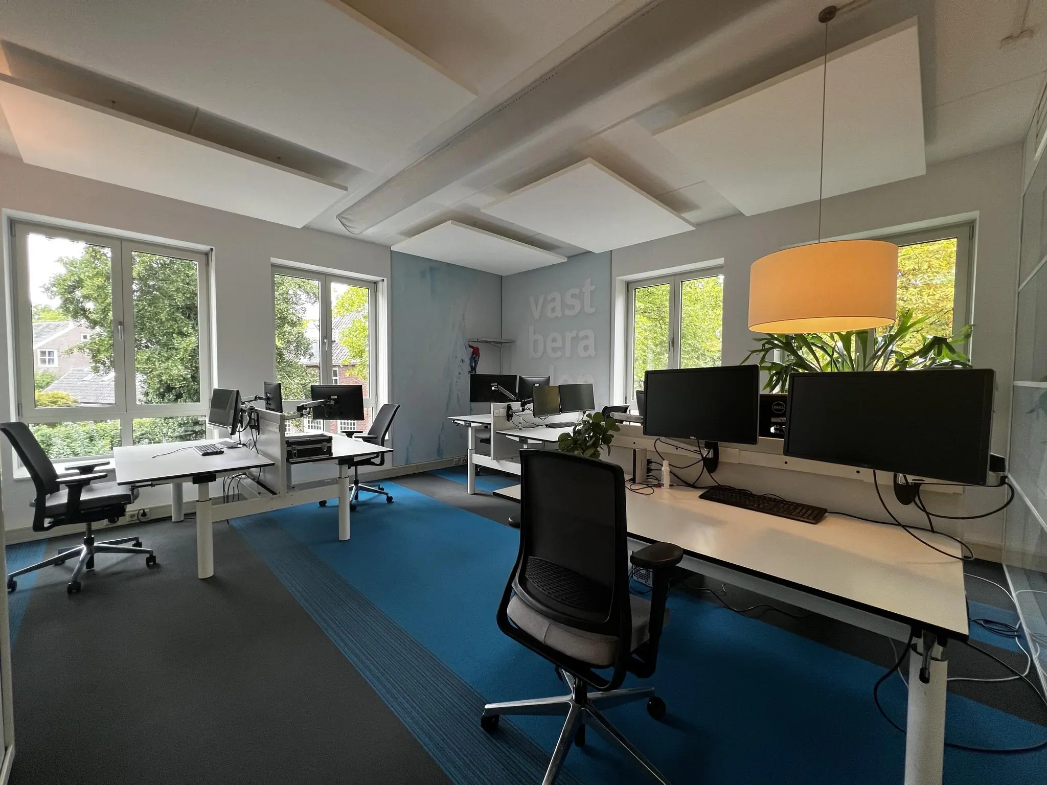Modern kantoor met meerdere werkplekken, grote ramen en de tekst "vastbera" op de muur aan de Driebergseweg.