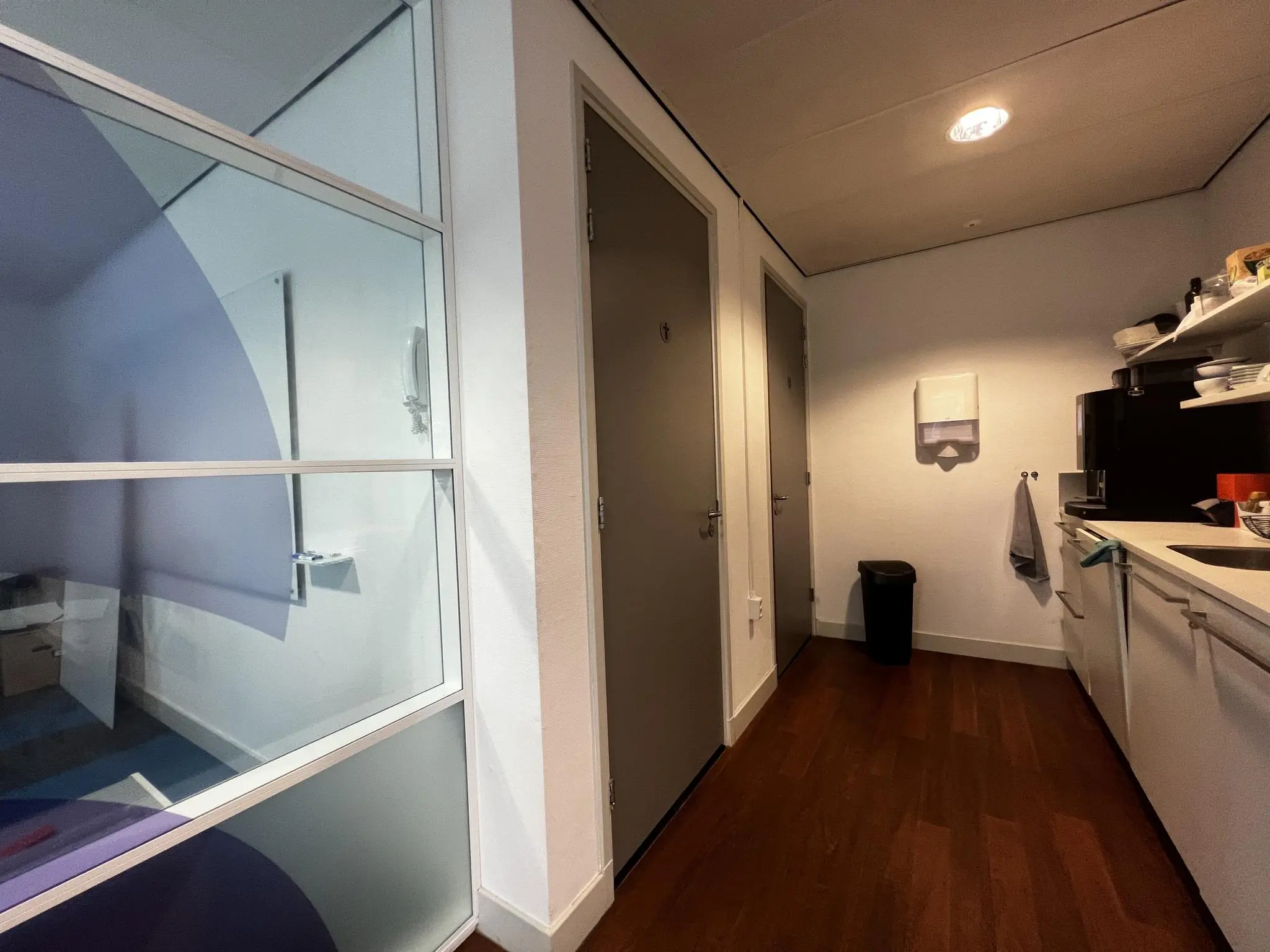 Keukenruimte met witte kasten, een donkerhouten vloer en twee grijze deuren in een kantoor aan de Driebergseweg.