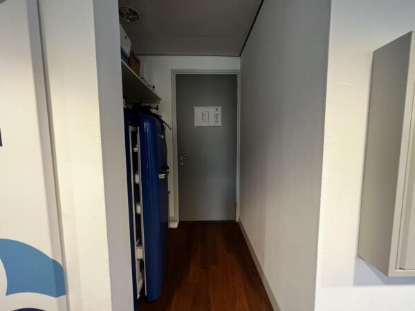 Gang met houten vloer, grijze deur en blauwe apparaten langs de linkermuur aan de Driebergseweg.