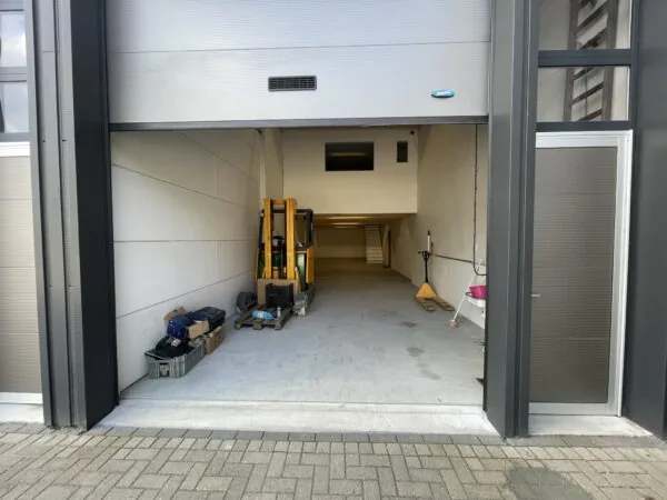 Openstaande garagedeur aan de Aalsmeerderweg met een magazijnruimte, palletwagen en gereedschap zichtbaar binnenin.