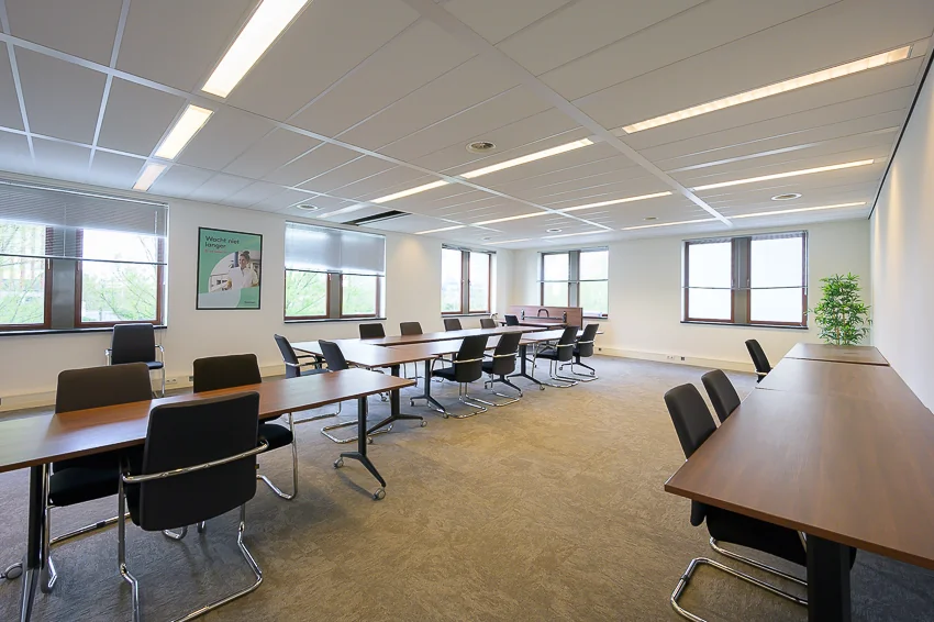 Vergaderruimte aan de Bahialaan met lange houten tafels, zwarte stoelen en grote ramen die natuurlijk licht binnenlaten.
