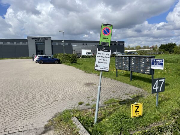 Vooraanzicht van het Storage Center Aalsmeer met parkeerplaats, brievenbussen en diverse borden, waaronder een te koop-bord en camerabewakingswaarschuwing.