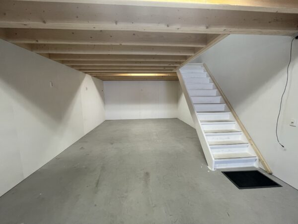 Een lege, strak afgewerkte kelderruimte met een betonnen vloer, houten plafondbalken en een witte trap aan de rechterzijde.