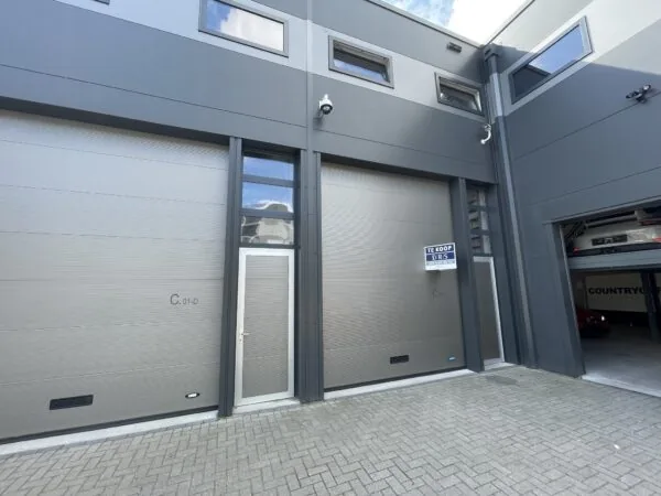 Bedrijfsunit aan de Aalsmeerderweg met grijze gevel, hoge garagedeur, loopdeur en een "Te Koop" bord van DRS makelaars.