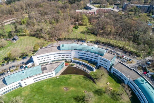 Luchtfoto van zorginstelling Sparrenheuvel, gelegen aan de rand van een bosrijk gebied met een gebogen, modern gebouw en omliggend groen.