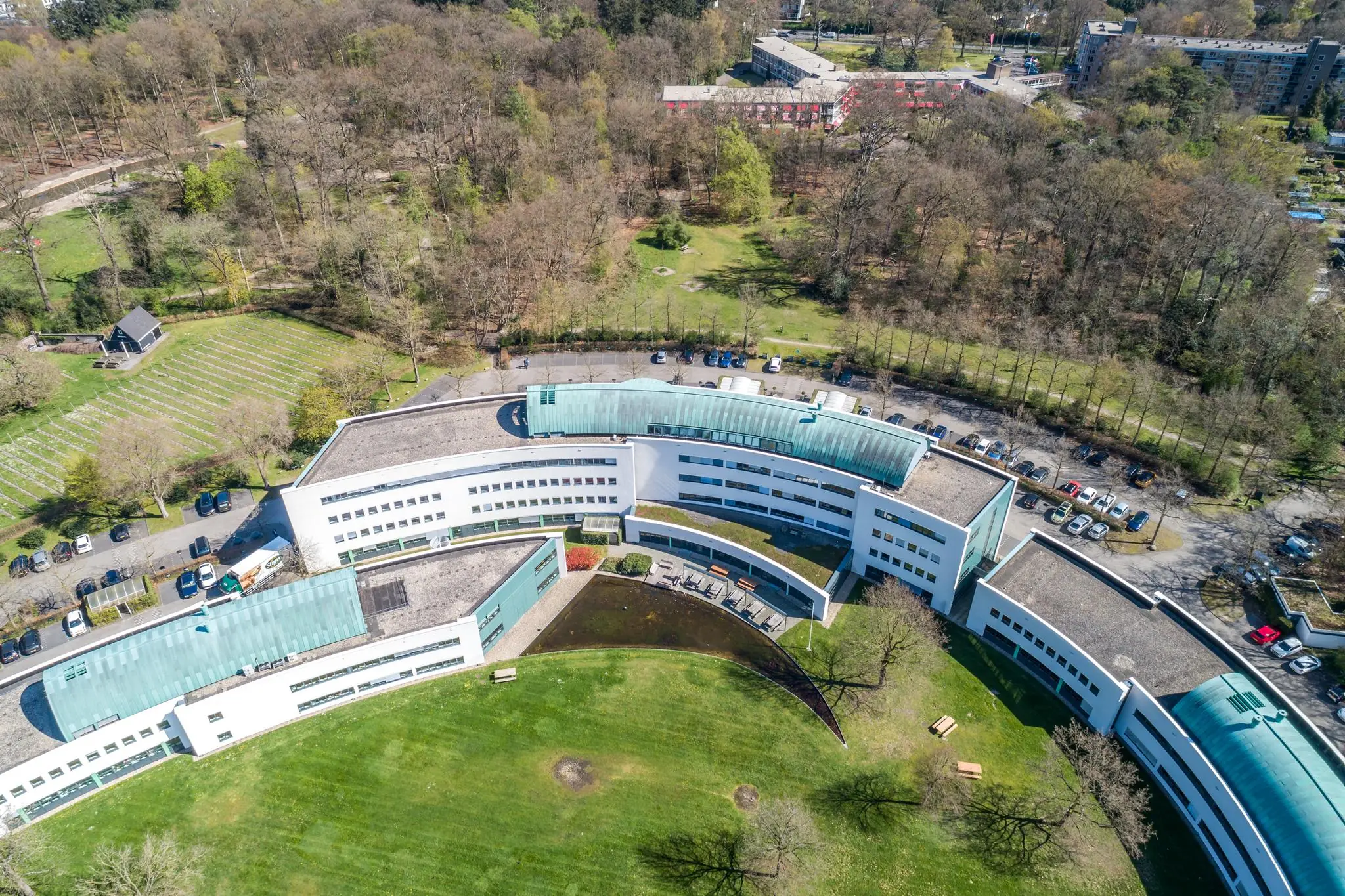 Luchtfoto van zorginstelling Sparrenheuvel, gelegen aan de rand van een bosrijk gebied met een gebogen, modern gebouw en omliggend groen.