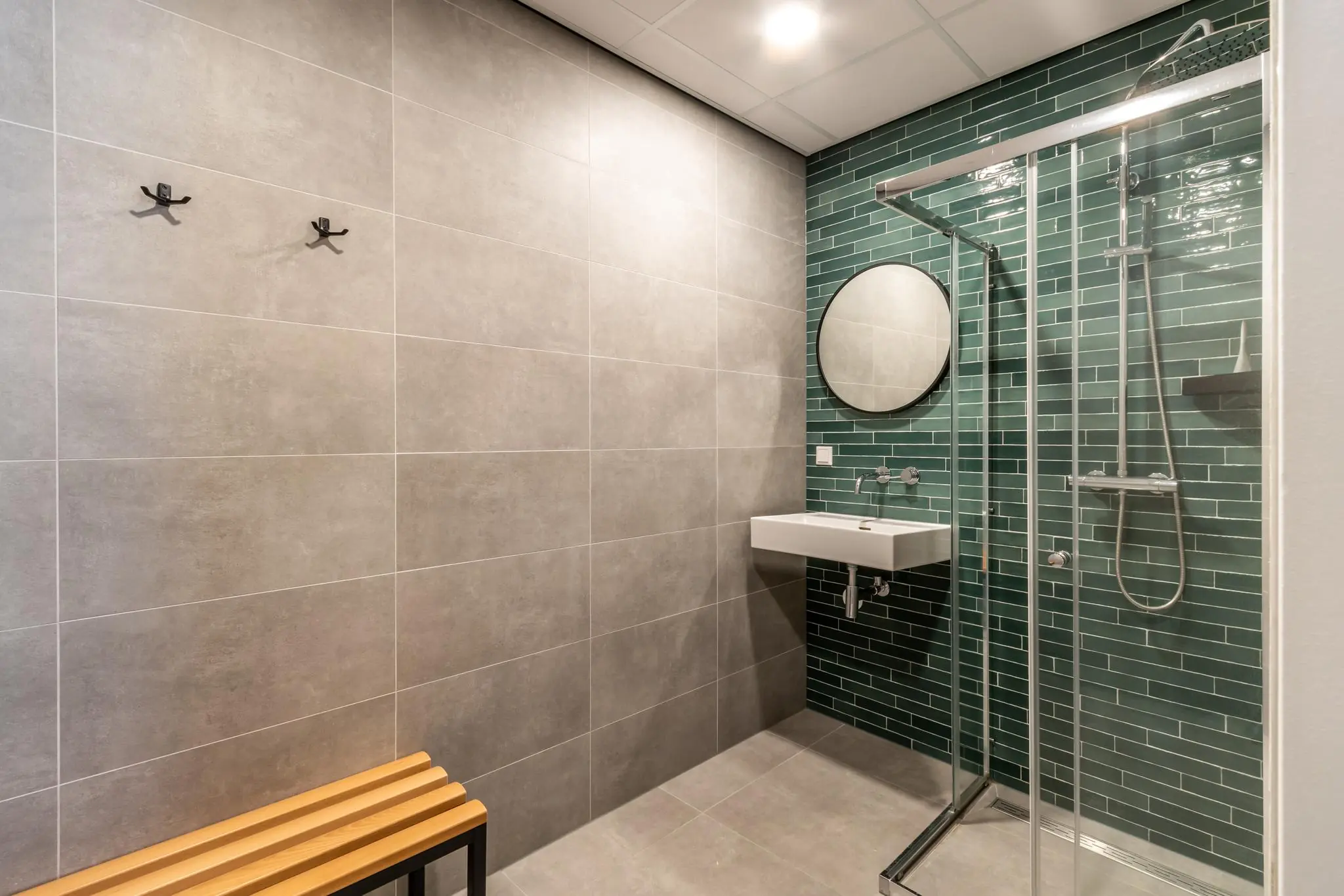 Moderne badkamer met een glazen douchecabine, groene wandtegels, ronde spiegel en een houten bankje.